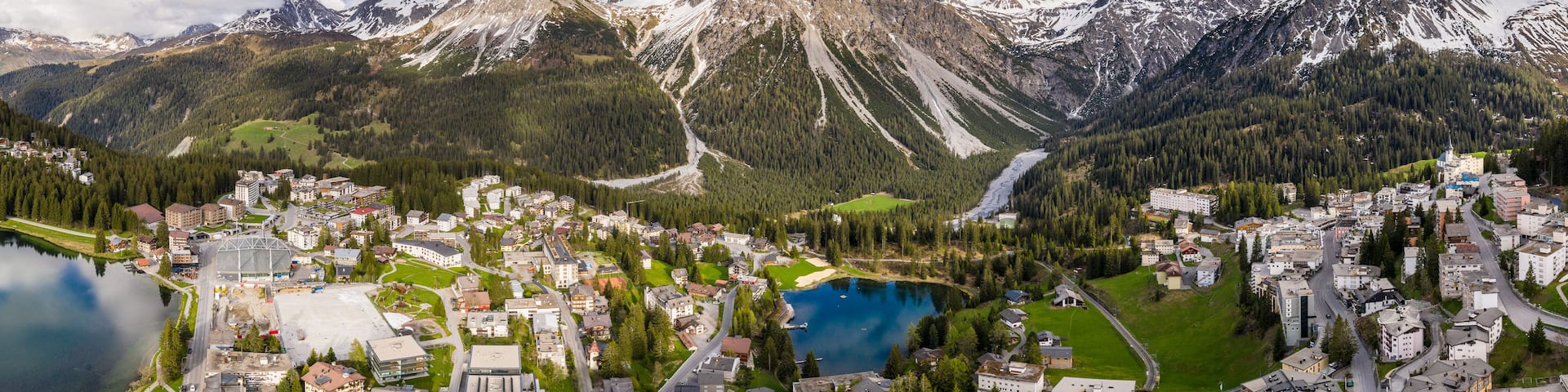 A mountainous landscape with a town nestled at the base, a lake, and snow-capped peaks in the background.
