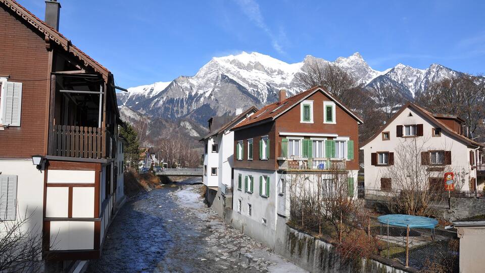 A snow-covered mountain range, a cobblestone street, and traditional European houses with green shutters.