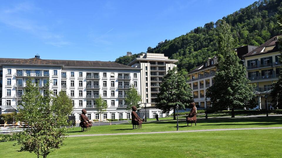 A large white building with multiple windows and balconies, surrounded by green grass and trees.