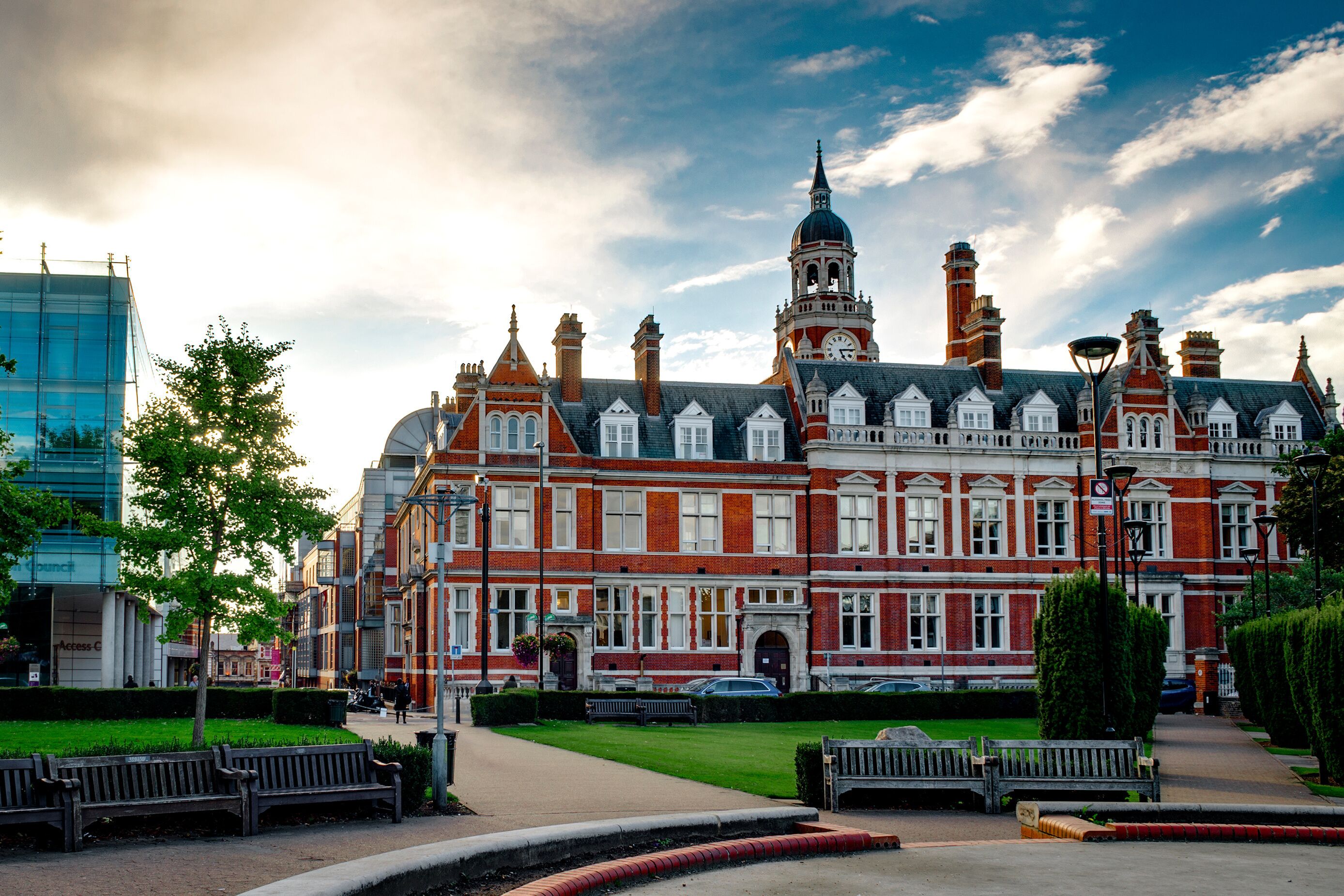 A historic red-brick building with a clock tower, surrounded by a well-maintained park with benches and a clear sky.
