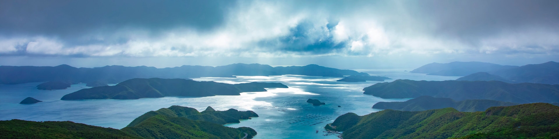 A coastal landscape with lush green hills, a bay with several islands, and a stormy sky.