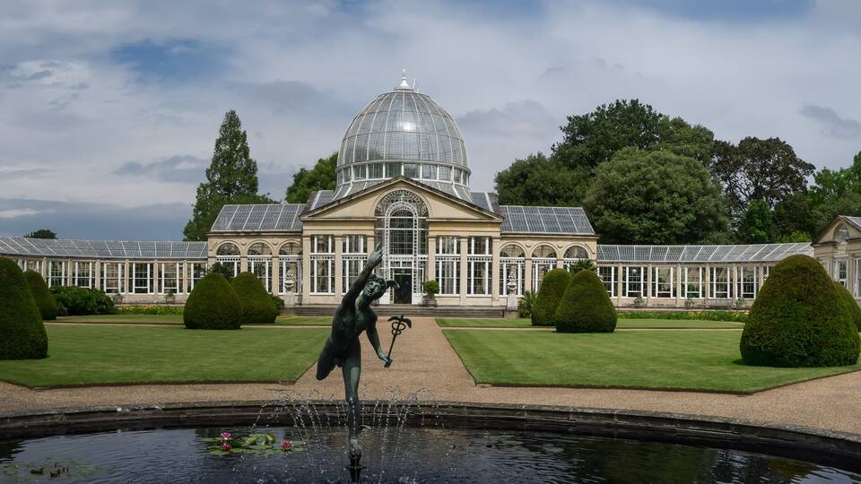 A large classical building with a central dome and symmetrical wings, surrounded by manicured gardens and a fountain.