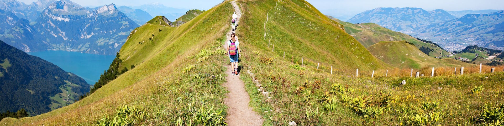 A hiking trail on a grassy mountain with a lake and distant mountains in the background.
