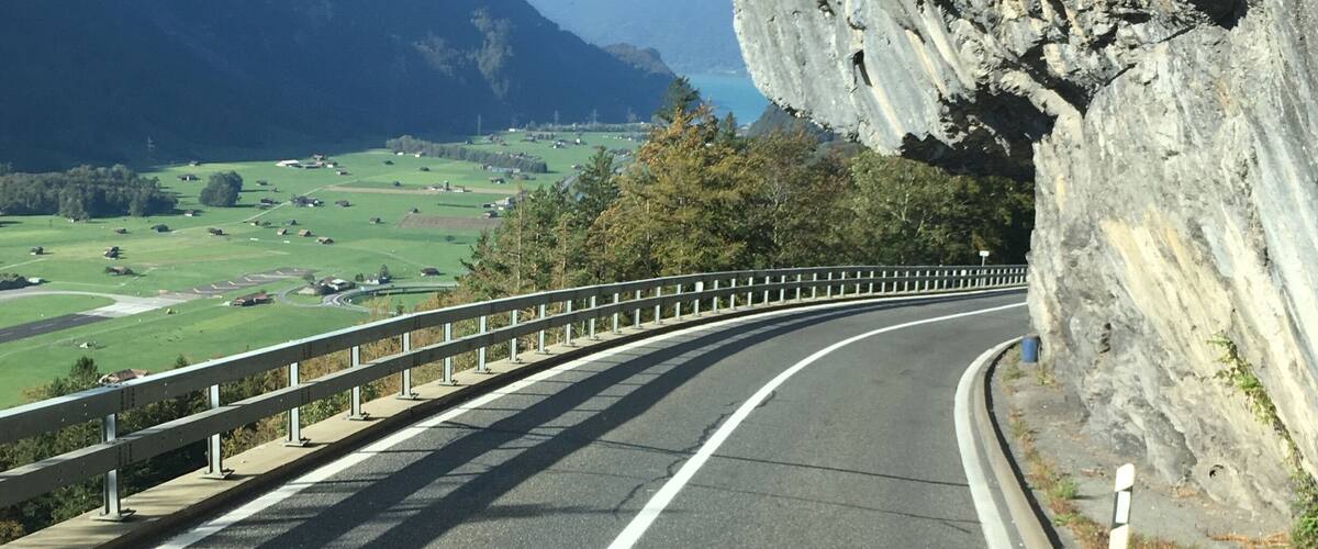 A winding mountain road with a guardrail, surrounded by lush green fields and mountains.