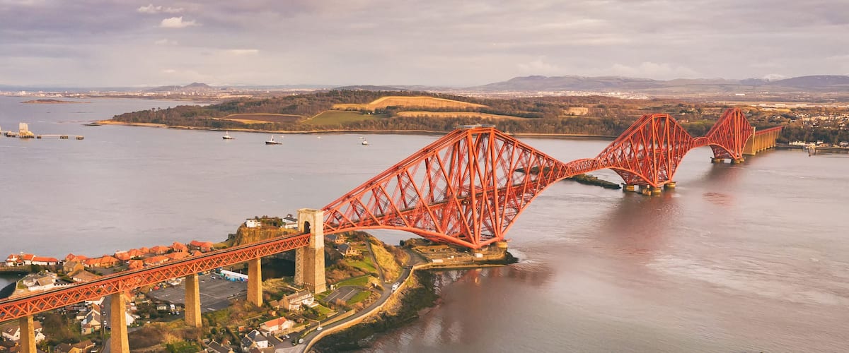 A red suspension bridge over a river with a town on the left and hills in the background.