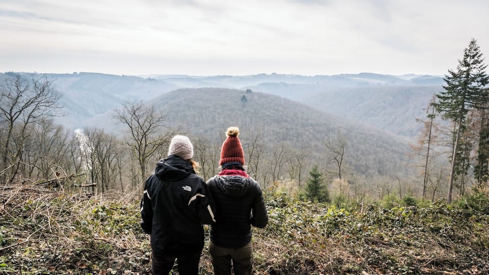 Two people standing on a hill overlooking a forested mountain range.