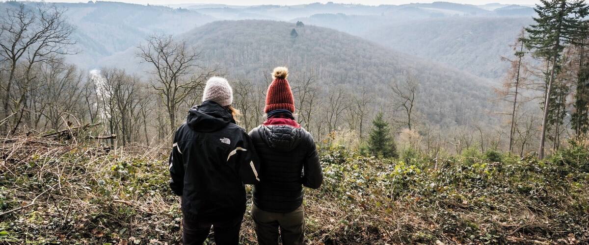 Two people standing on a hill overlooking a forested mountain range.