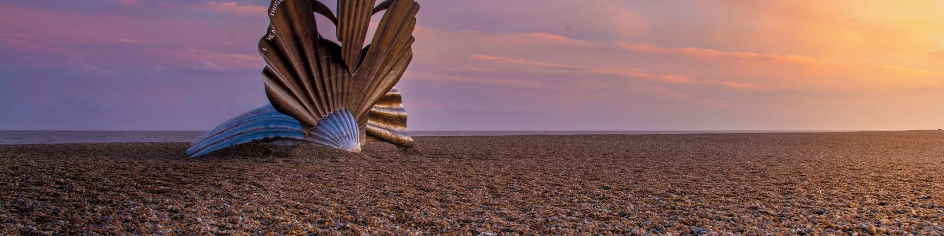 A large metallic sculpture resembling a shell on a pebbled beach at sunset.