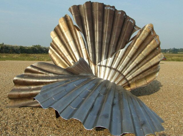 A large metallic sculpture resembling a scallop shell on a gravel surface.