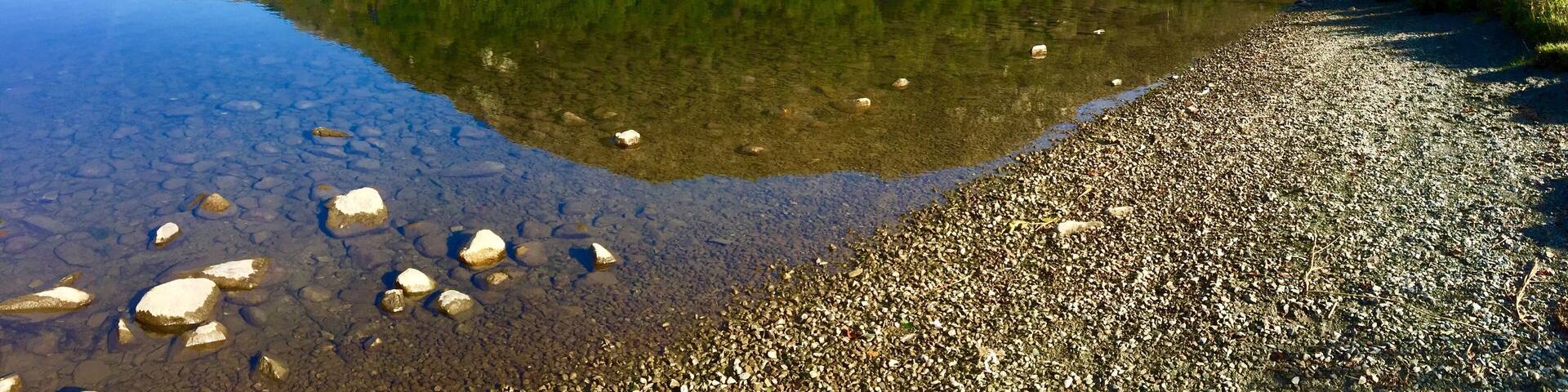 A calm lake with clear reflections, surrounded by a forested mountain and a pebbly shore.