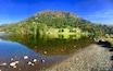 A calm lake with clear reflections, surrounded by a forested mountain and a pebbly shore.