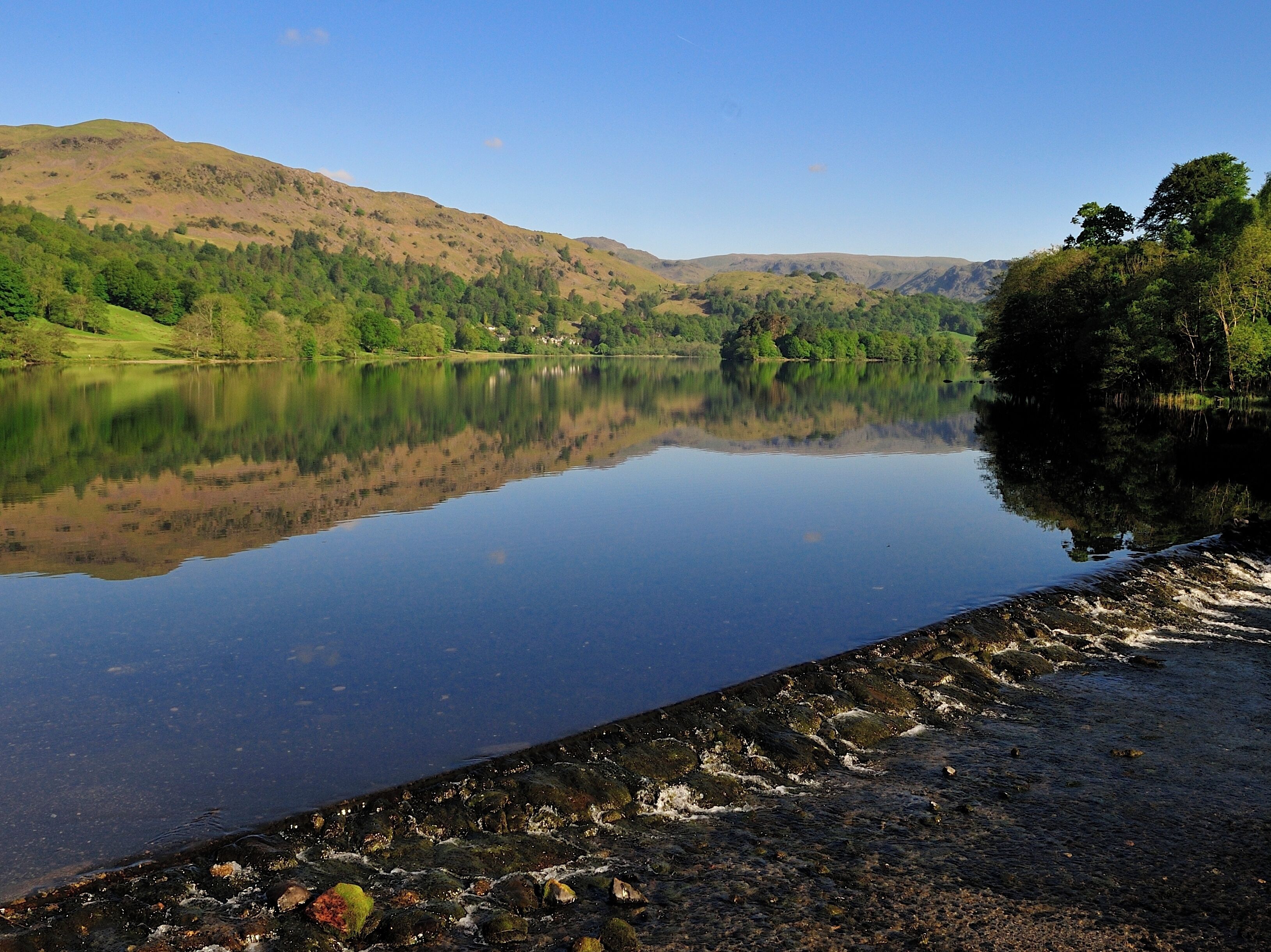 A calm lake with clear reflections, surrounded by green hills and trees.