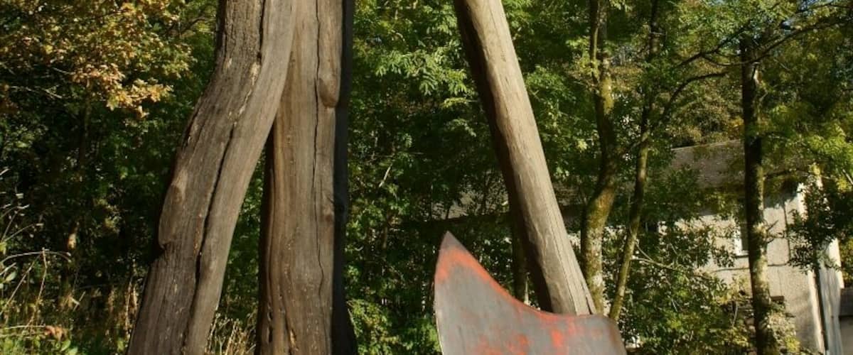 A wooden sculpture of two figures holding a large red object, set in a stone pathway with trees in the background.