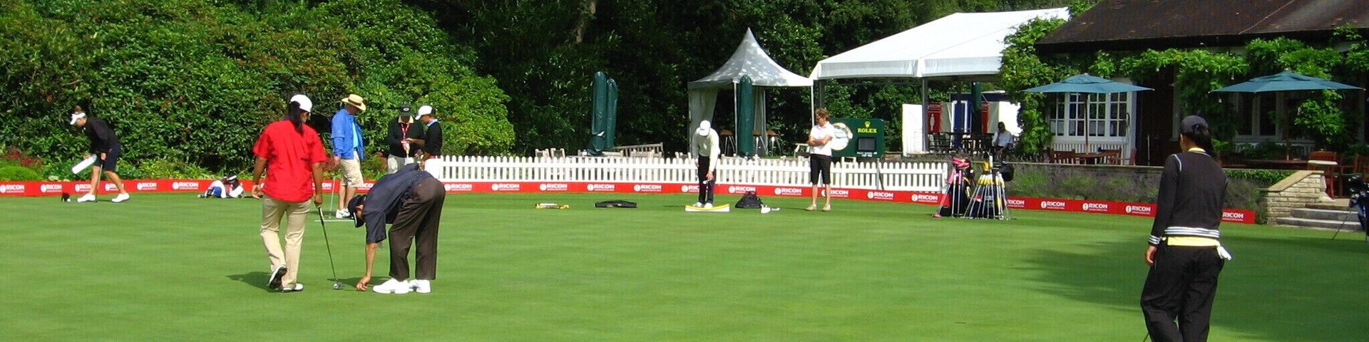 Un campo de golf con una cerca blanca de piquetes, una carpa y árboles al fondo.