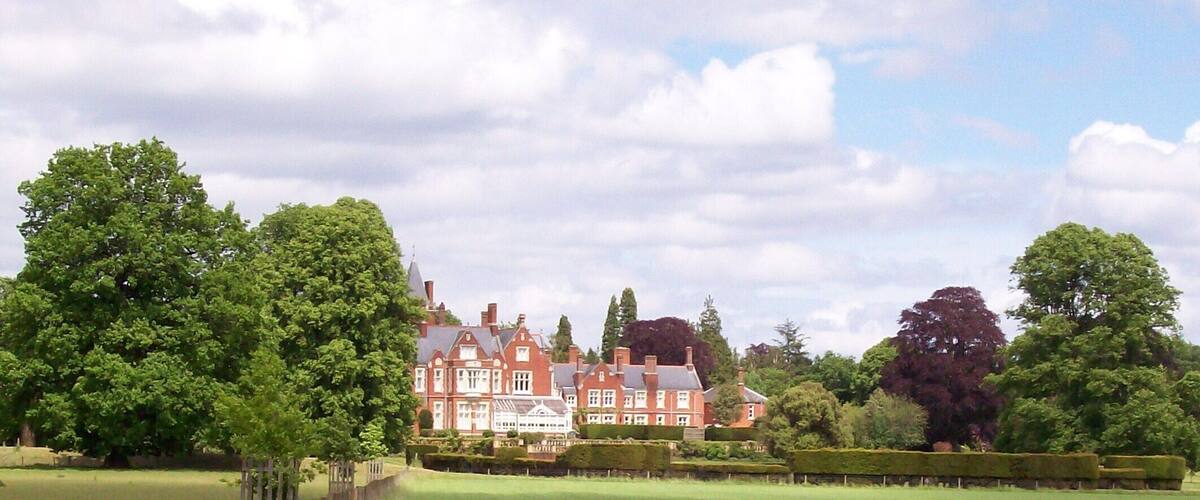 A large, historic red building surrounded by lush green trees and manicured hedges.