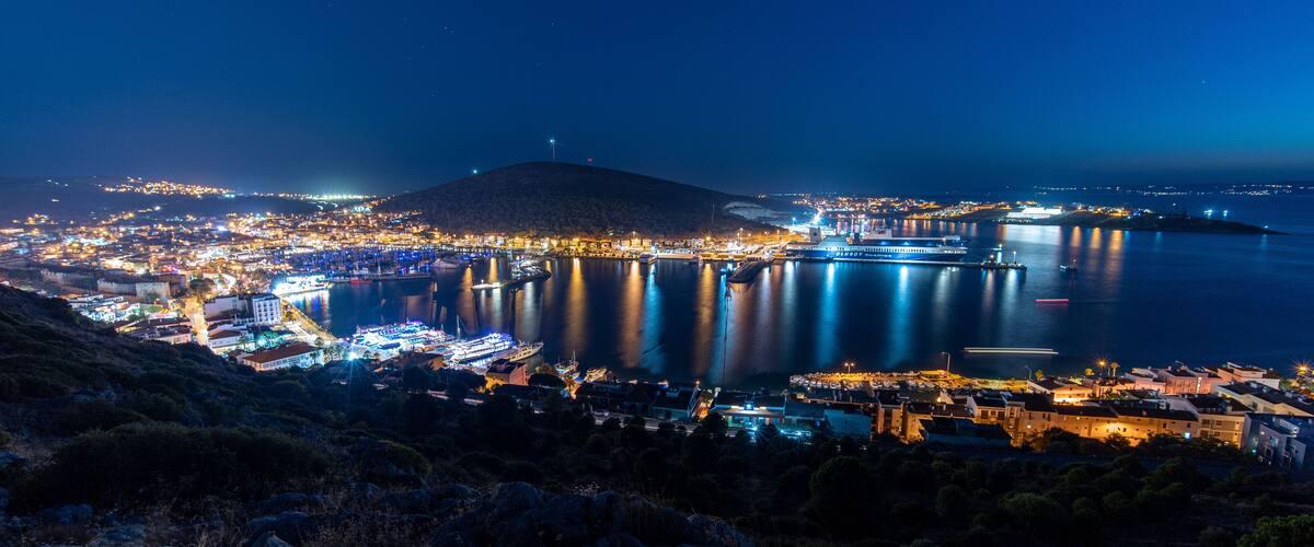 A coastal city at night with a prominent hill, a harbor with boats, and a distant island.