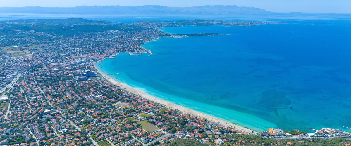 Luchtbeeld van een kustplaats met een lang strand, dichtbebouwde woonwijken en een heldere blauwe zee.