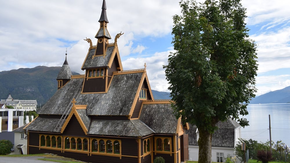 A wooden church with a pointed steeple and a large tree in front.