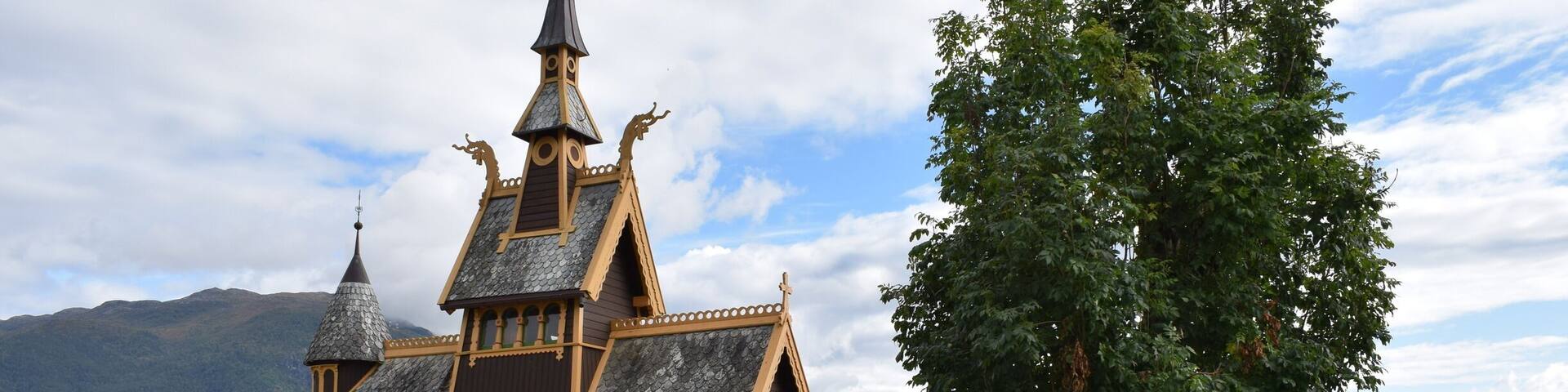 A wooden church with a pointed steeple and a large tree in front.