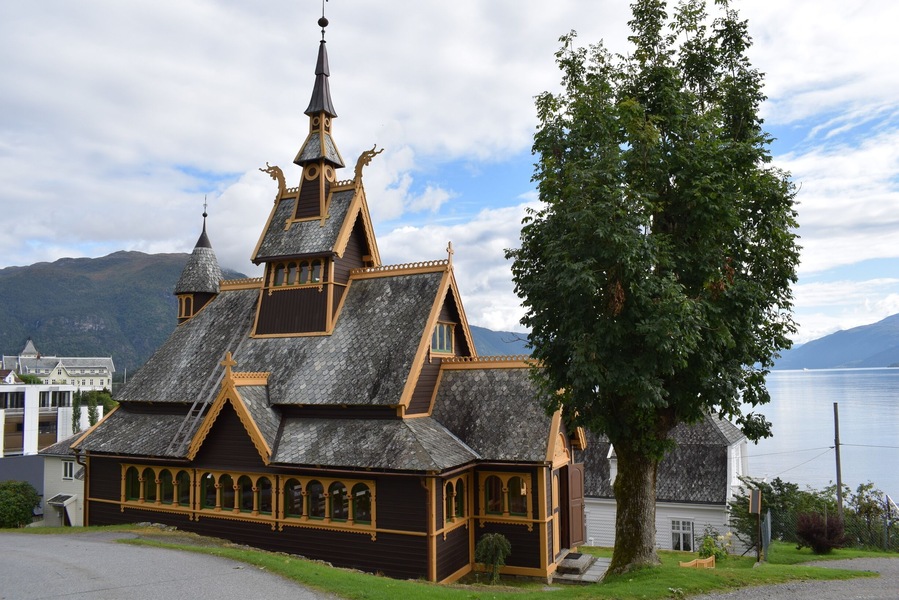 Una iglesia de madera con un campanario puntiagudo y un árbol grande en primer plano.