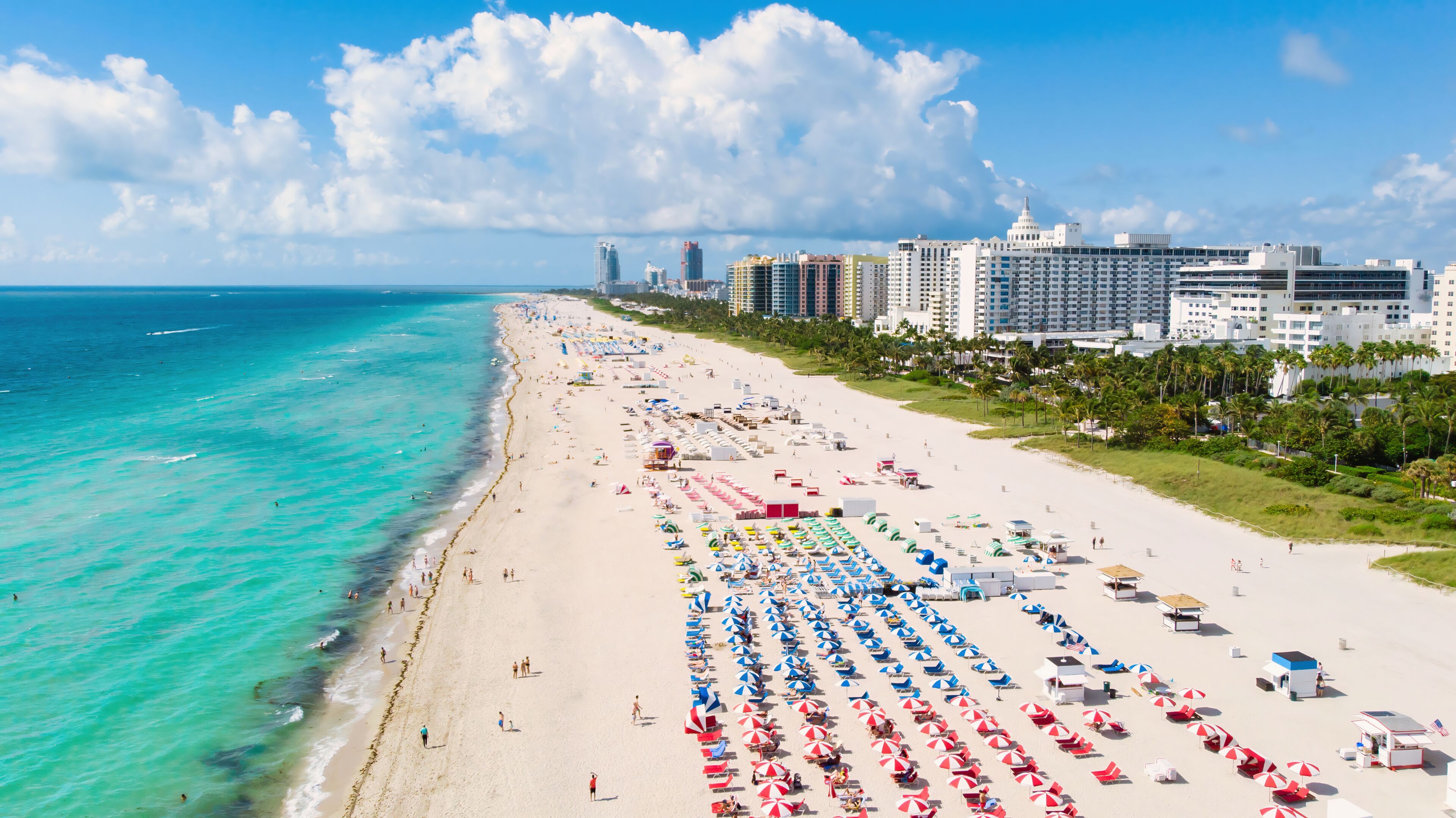 Ein Strand mit Reihen von Sonnenschirmen und Liegestühlen, einem klaren blauen Ozean und einer Stadtlandschaft im Hintergrund.