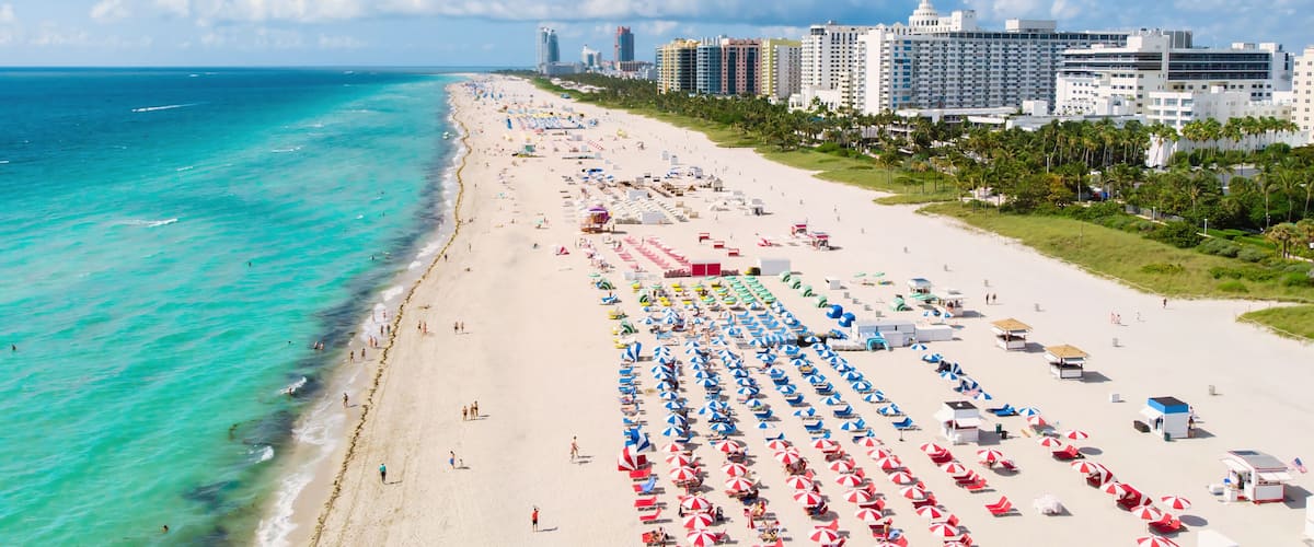 Une plage bordée de rangées de parasols et de chaises longues, une mer bleue et limpide, et, au loin, le profil d’une ville.