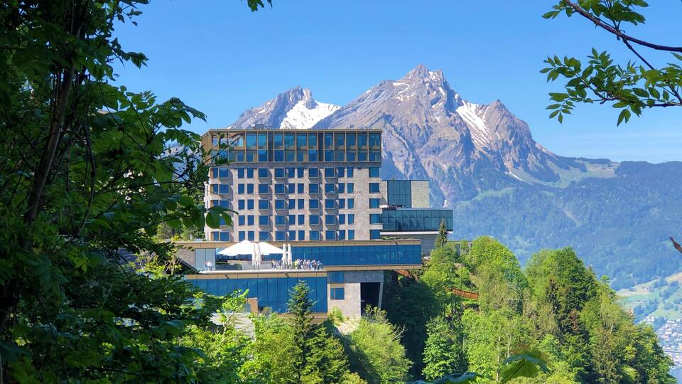 A modern building with a glass facade, surrounded by lush greenery and a mountain range in the background.