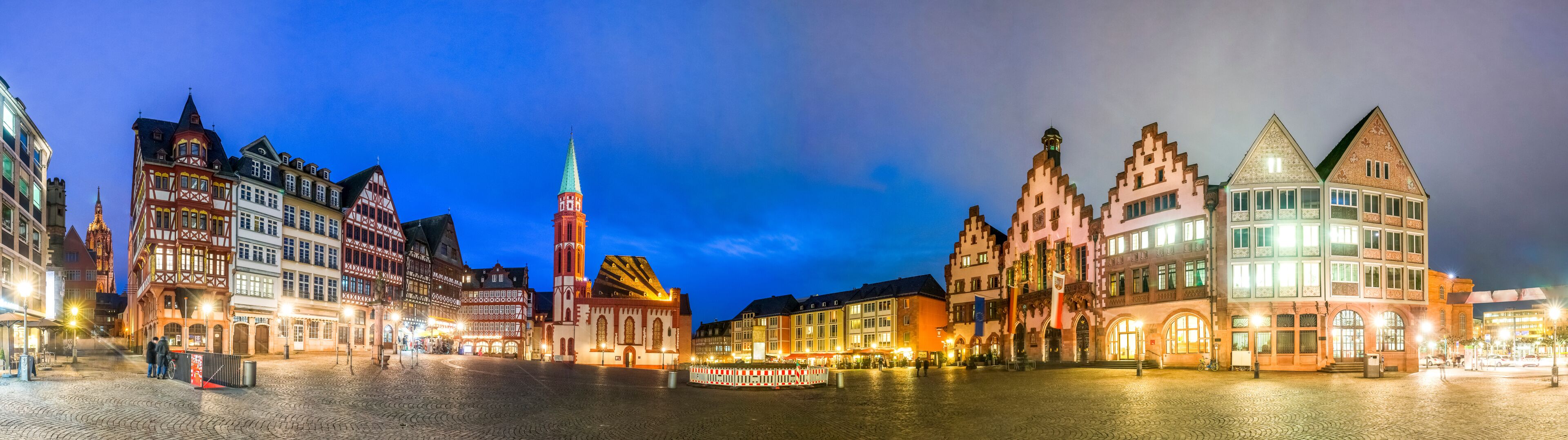 Une place historique au cœur d’une ville européenne, au crépuscule, avec ses bâtiments traditionnels à colombages et son imposant clocher d’église.