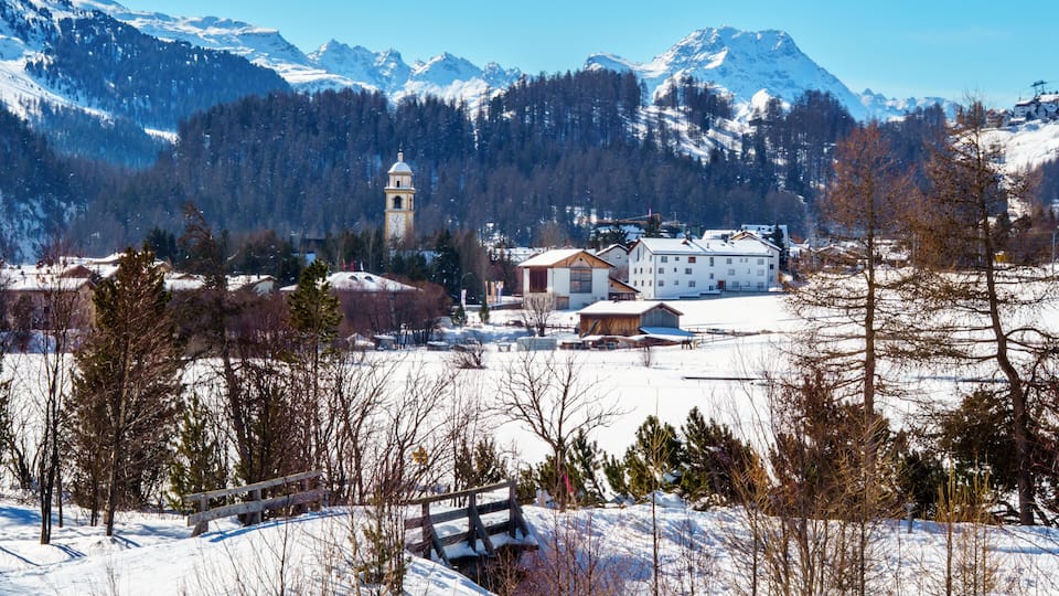 A snow-covered village with a church tower, buildings, and mountains in the background.