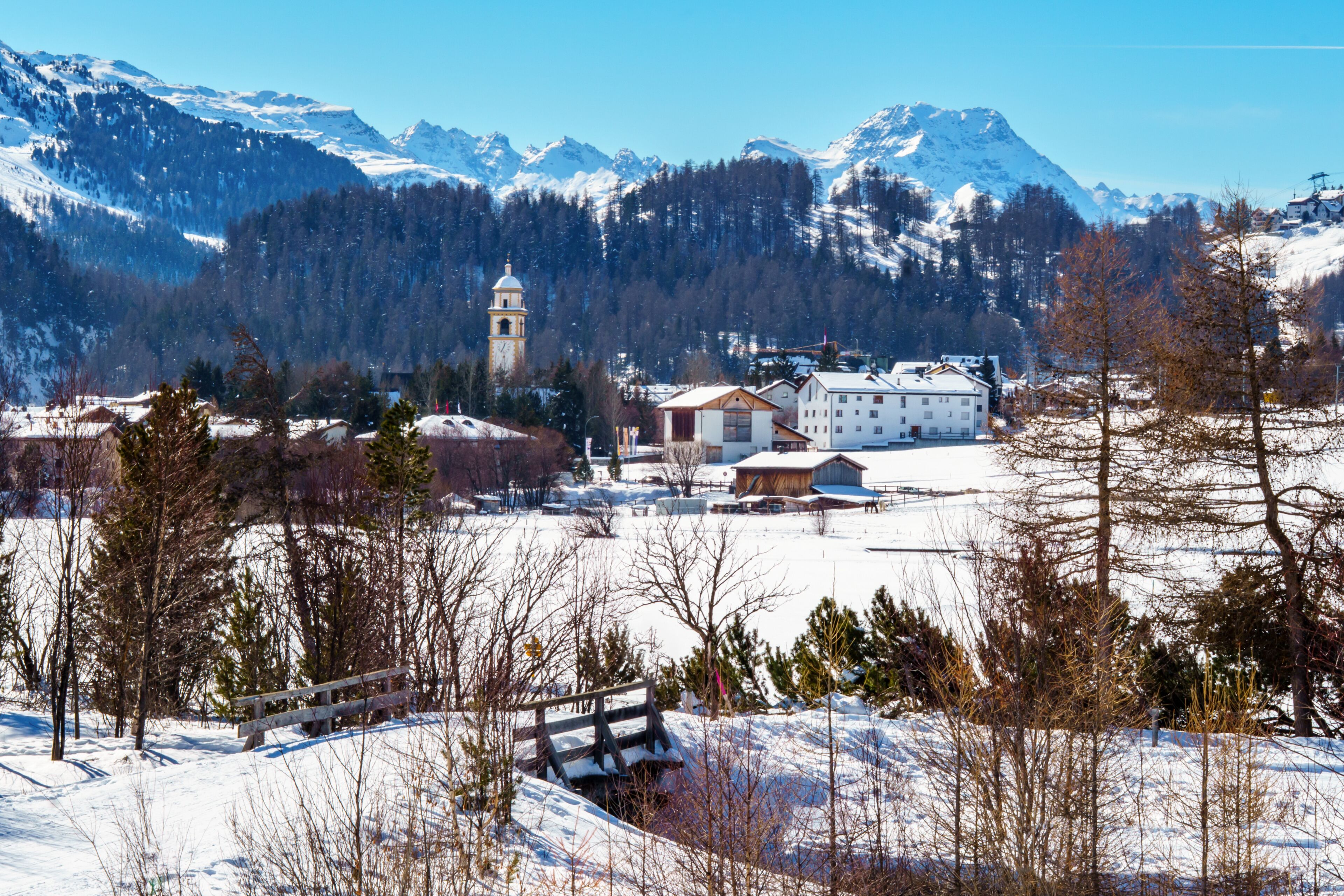 A snow-covered village with a church tower, buildings, and mountains in the background.