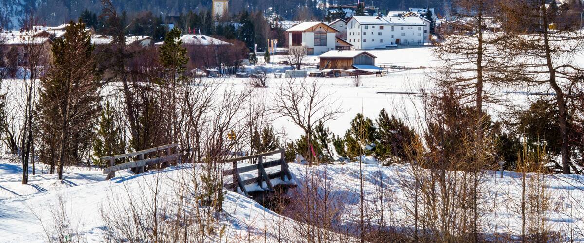 A snow-covered village with a church tower, buildings, and mountains in the background.