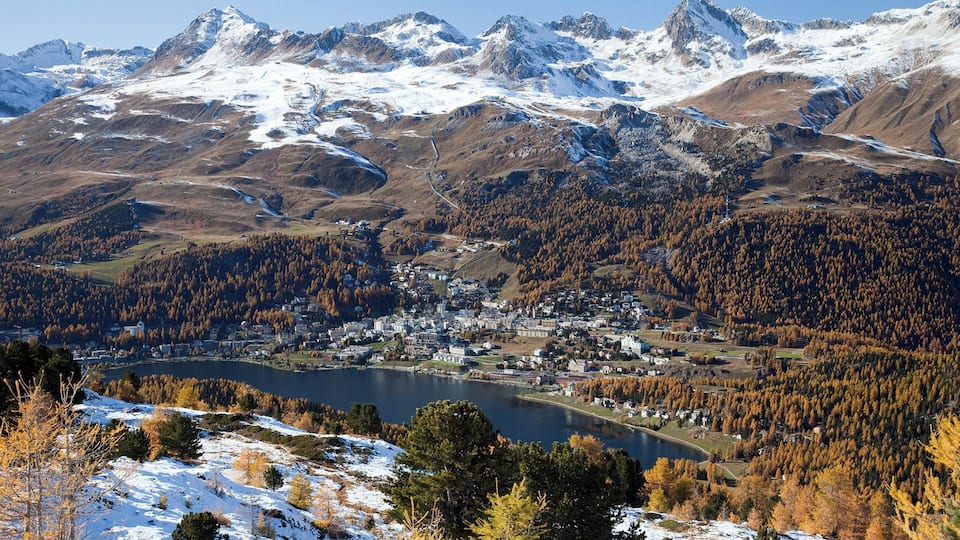 A mountainous landscape with a town nestled in a valley, surrounded by snow-capped peaks and autumn-colored forests.