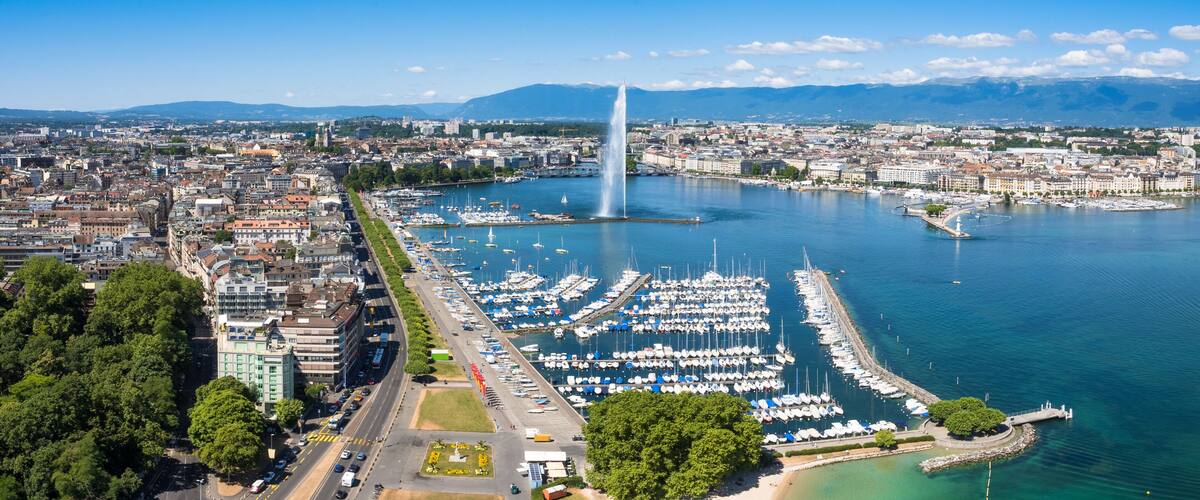 A marina with numerous boats, a prominent fountain, and a cityscape with mountains in the background.