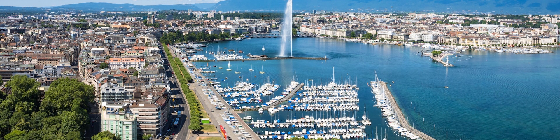 A marina with numerous boats, a prominent fountain, and a cityscape with mountains in the background.