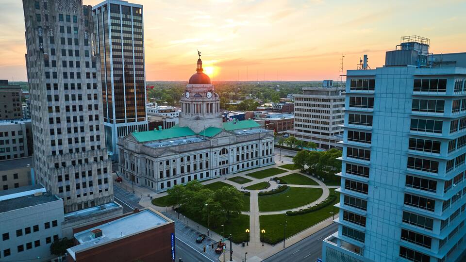 A cityscape with a prominent building featuring a dome and clock tower, surrounded by modern high-rise buildings.