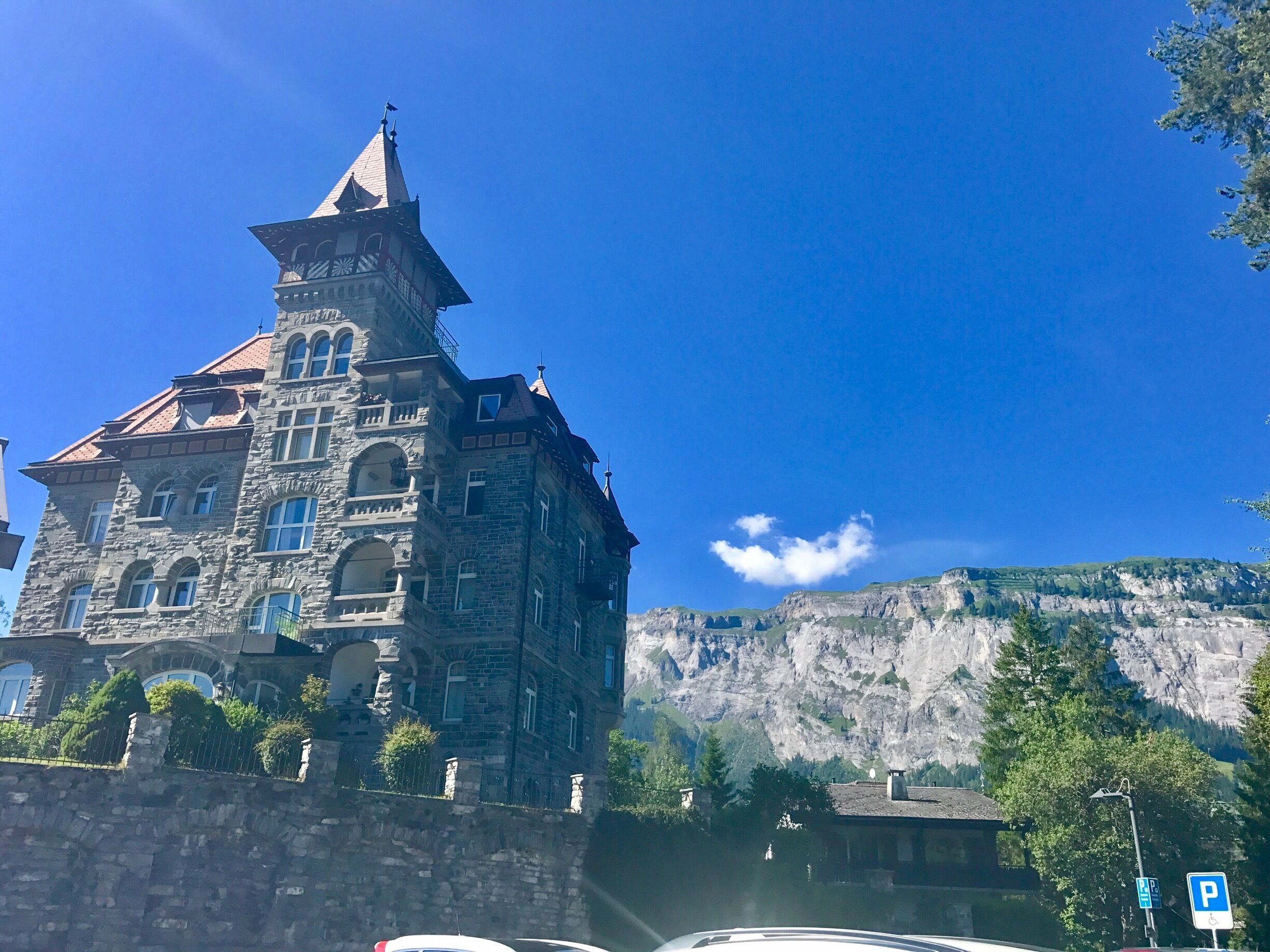 A historic stone building with a steep roof and a tower, set against a backdrop of a mountainous landscape.