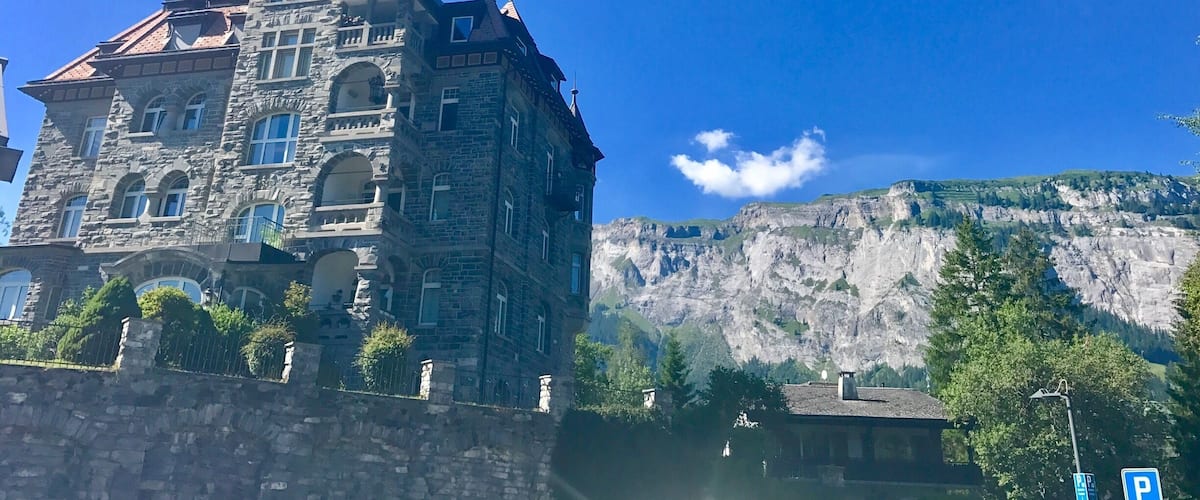 A historic stone building with a steep roof and a tower, set against a backdrop of a mountainous landscape.