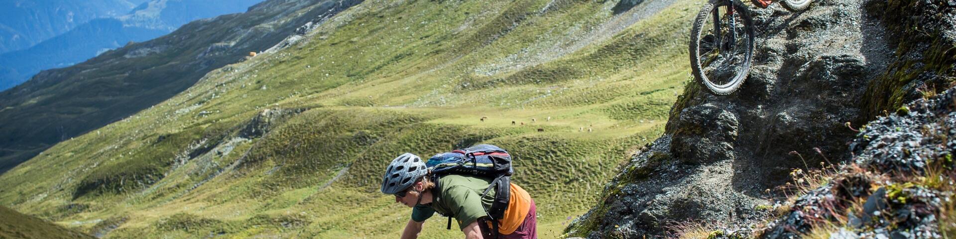 Two mountain bikers on a rocky trail with a mountainous landscape in the background.