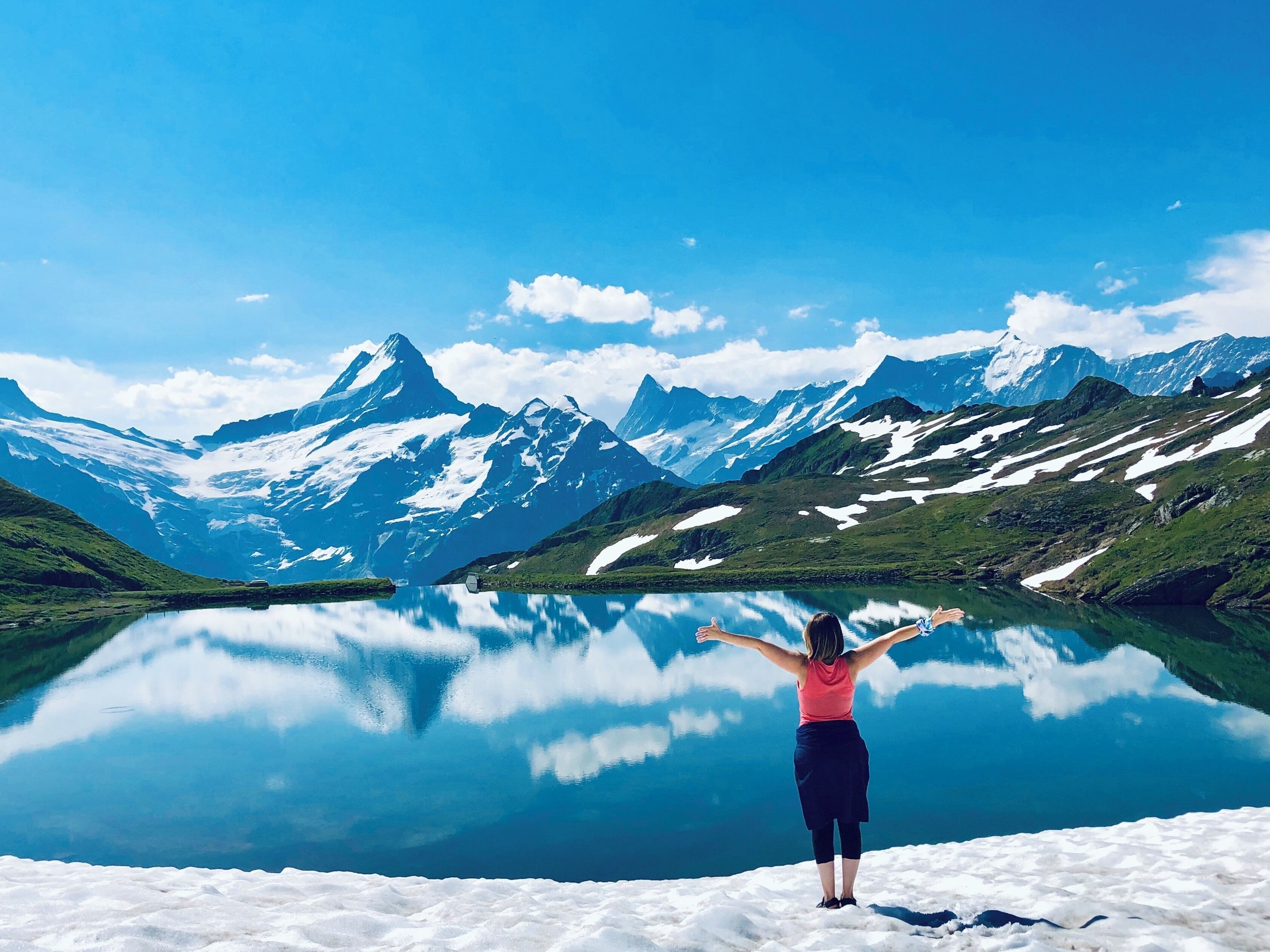 Una persona de pie sobre una superficie nevada, con los brazos extendidos, contemplando un lago tranquilo y montañas cubiertas de nieve.