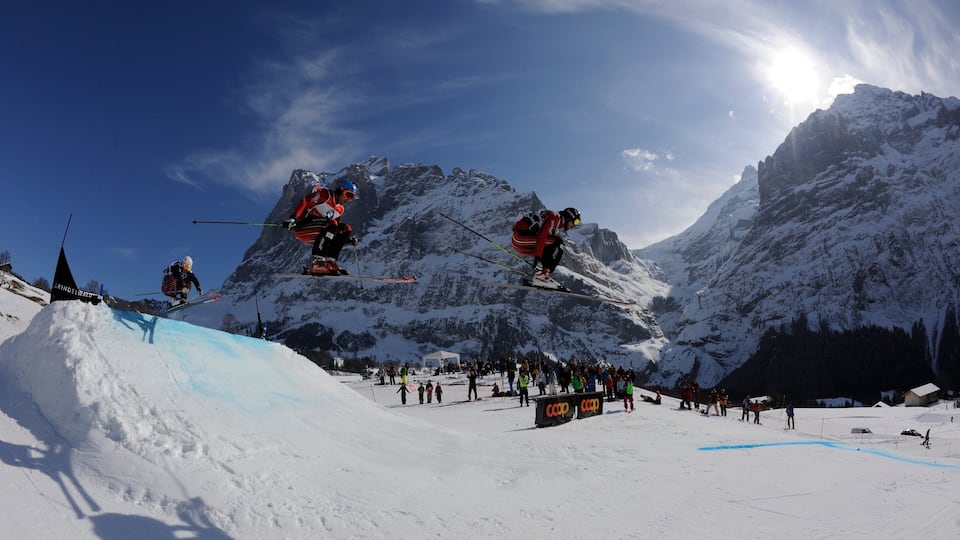 A ski jump with skiers in mid-air against a mountainous backdrop.