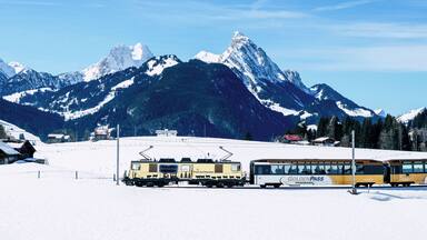 A train station with a yellow train and a bus labeled "Golden Pass Panorama" in a snowy mountainous area.