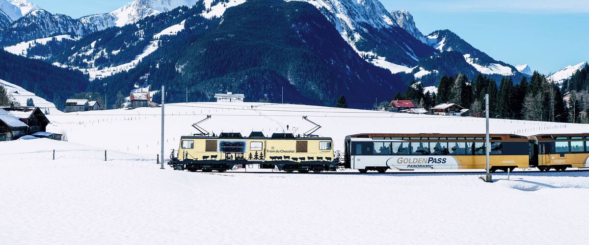 A train station with a yellow train and a bus labeled "Golden Pass Panorama" in a snowy mountainous area.