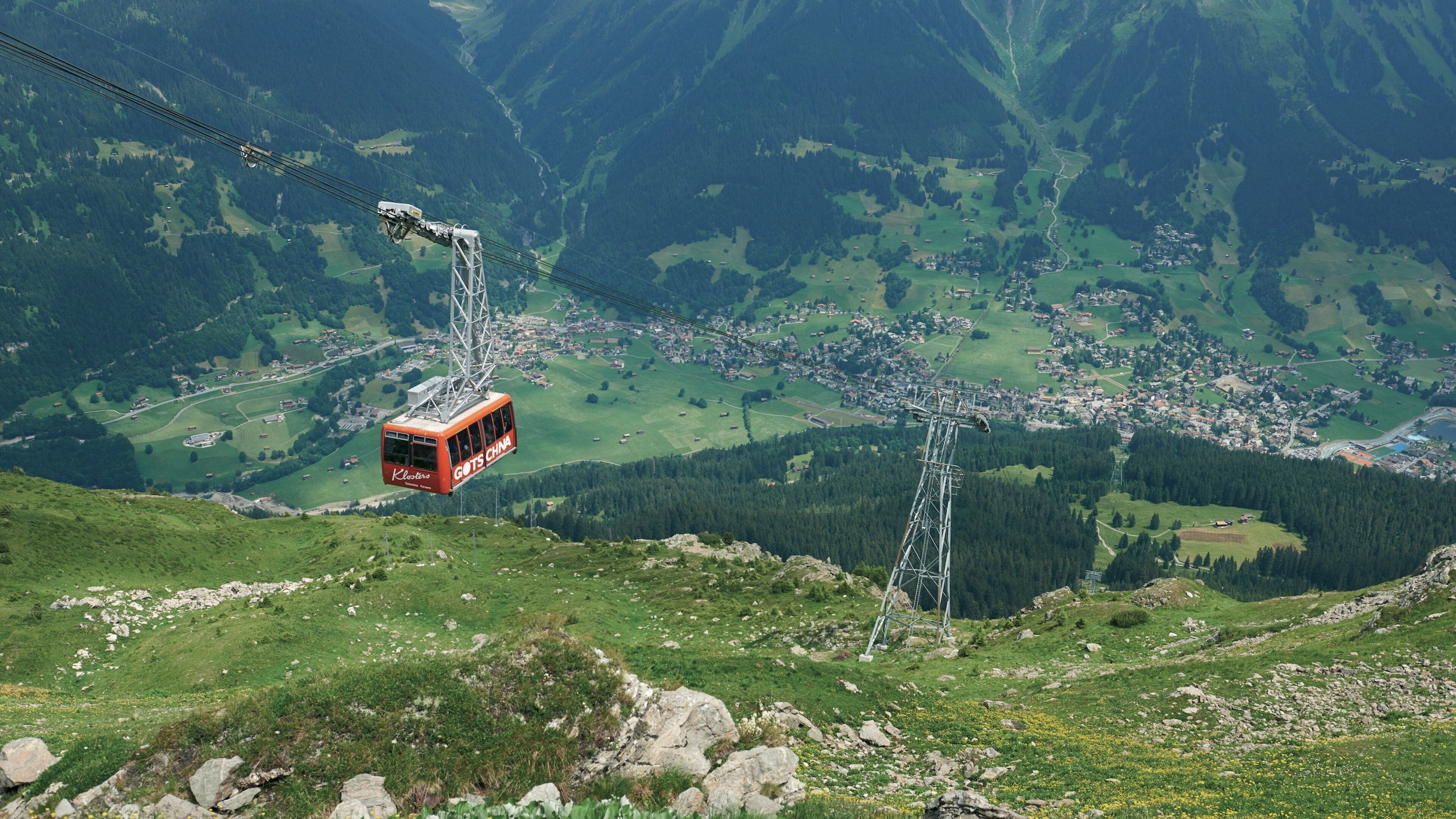 A cable car over a mountainous landscape with green valleys and scattered buildings.