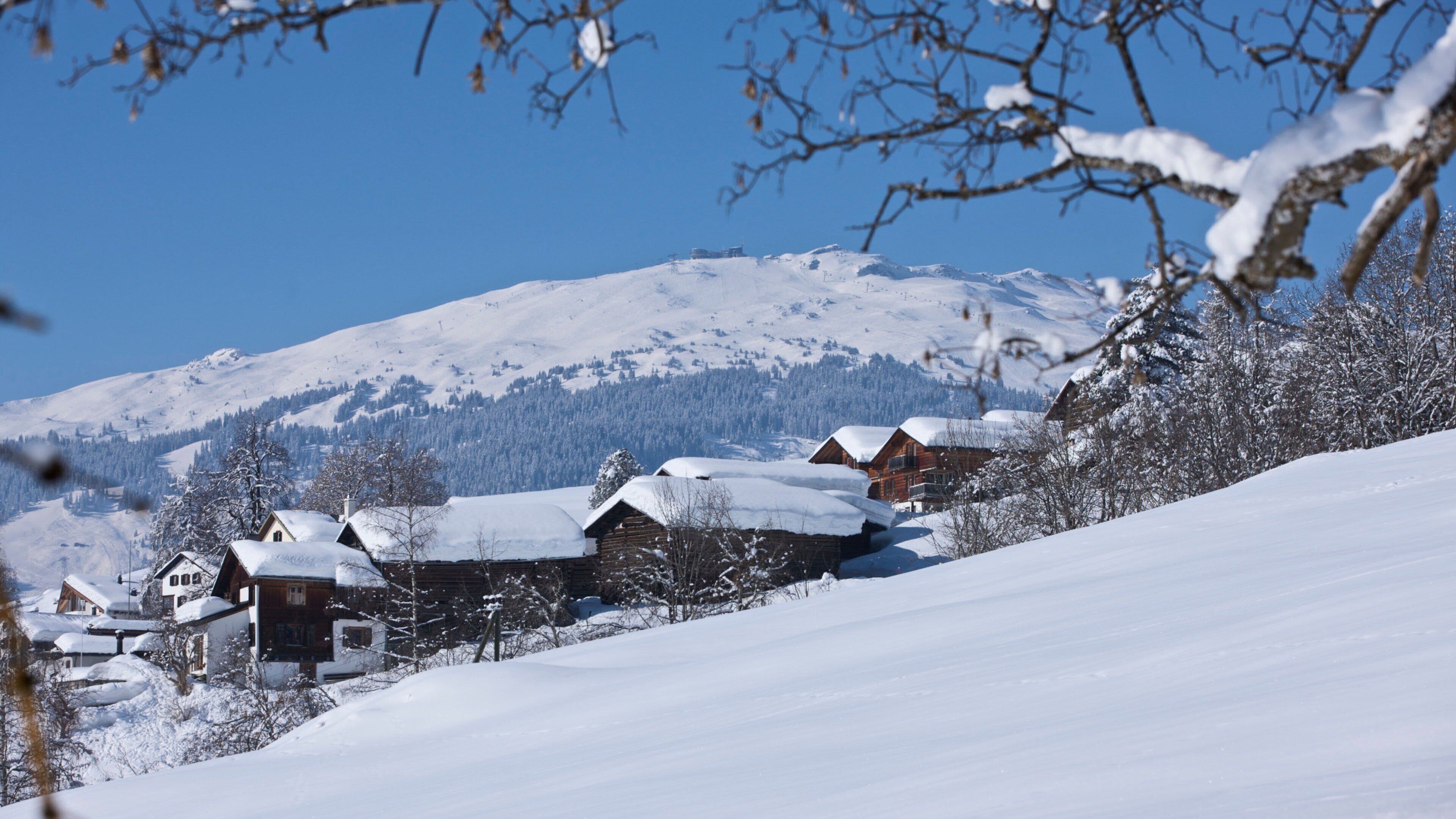 Ein mit Schnee bedeckter Berg unter einem klaren, blauen Himmel. Holzhäuser mit Schnee auf den Dächern sind zu sehen.