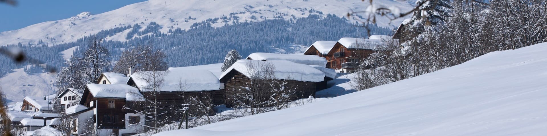 A snow-covered mountain with a clear blue sky. There are wooden houses with snow on their roofs.