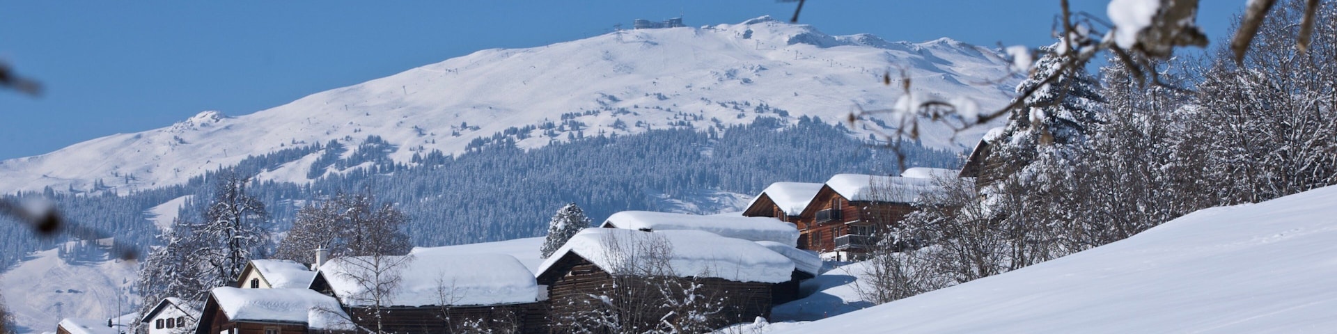 Montanha coberta de neve com céu azul límpido. Há casas de madeira com neve nos telhados.