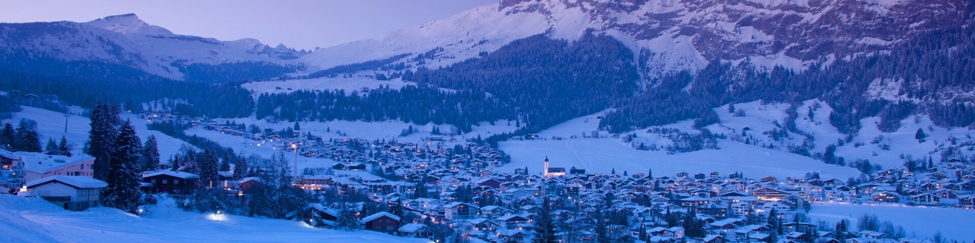 A snow-covered mountain town at dusk with illuminated buildings and a clear view of the surrounding peaks.