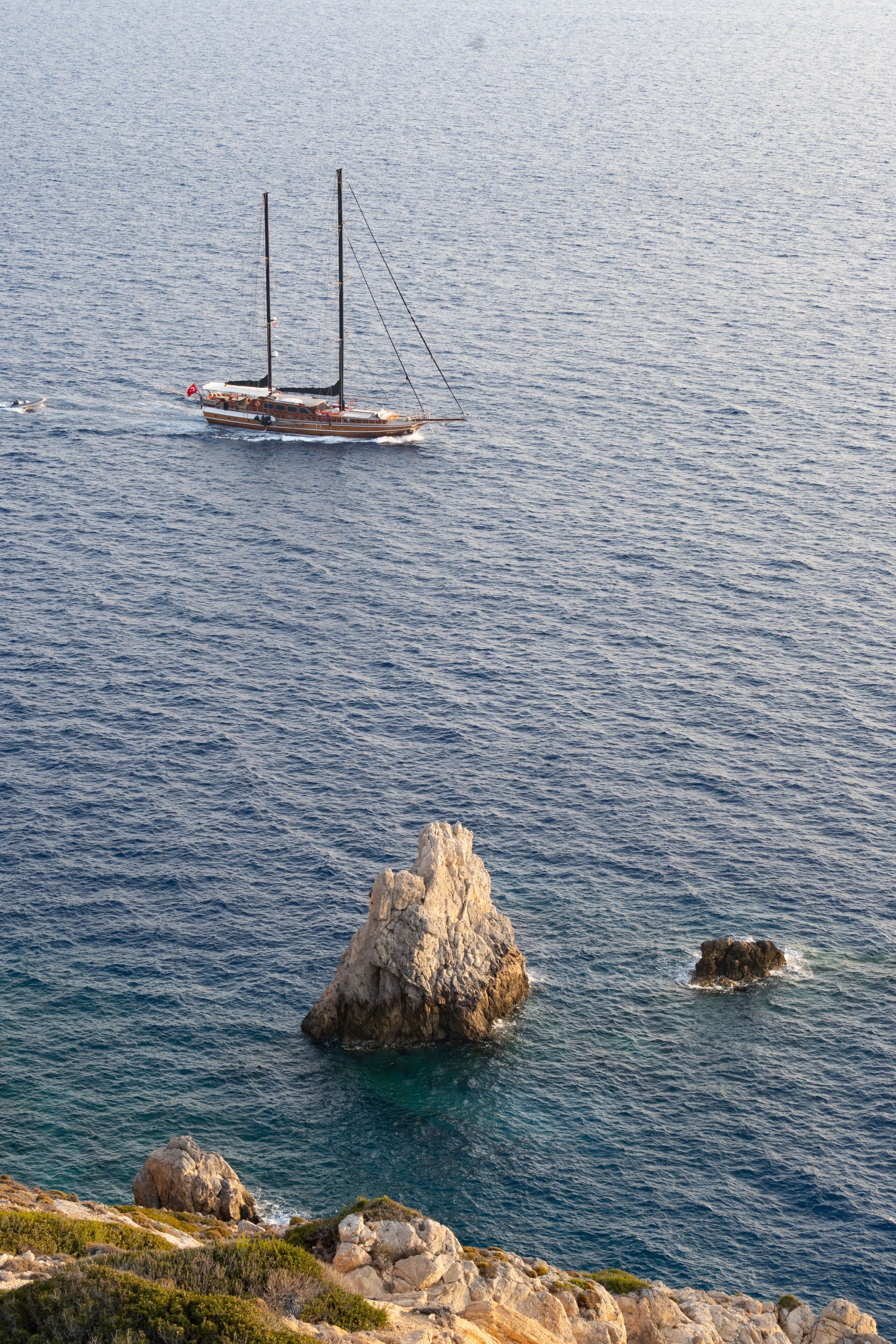 A sailboat on calm sea waters, rocky coastline, and a prominent rock formation.