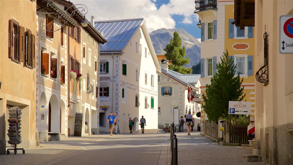A cobblestone street lined with traditional European buildings, a mountain in the background, and people walking.