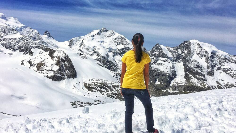 Une personne se tient sur une montagne enneigée, avec un terrain rocheux et des sommets enneigés en arrière-plan.